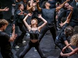 A group of actors in black clothing stretch in a variety of ways.