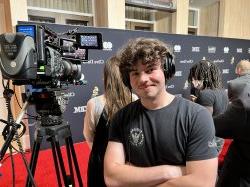A student stands next to a camera and wears headphones with a red carpet in the background.