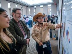 A student wearing a cowboy hat reviews his poster as two people look on.