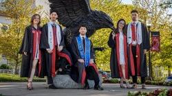 The Povolo quintuplets in graduation gowns stand in front of a large Red Hawk statue.