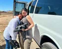 A student holding a video camera hugs another student in the desert.